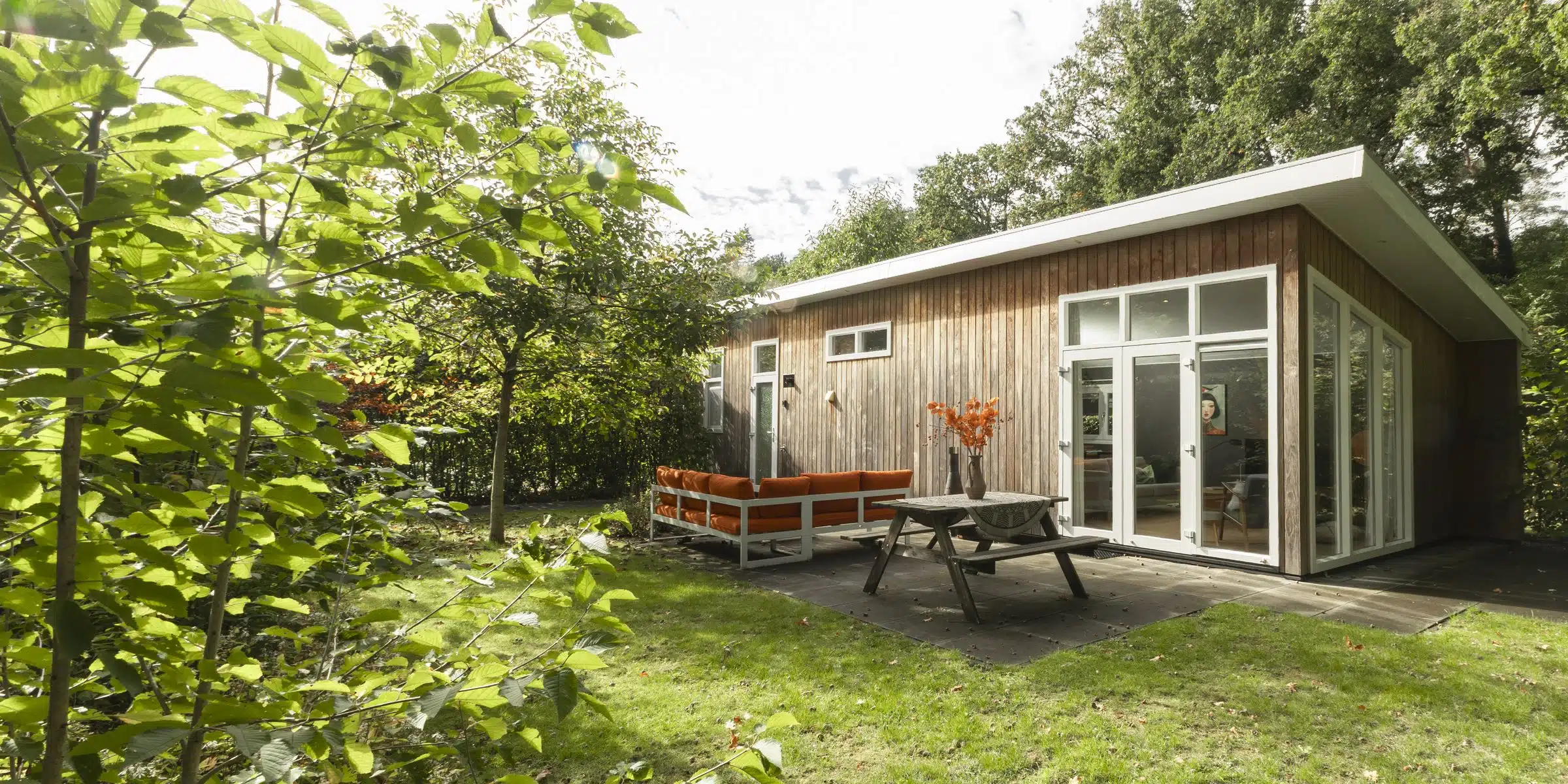 Houten vakantiehuis met terras, oranje loungebank en picknicktafel in een bosrijke tuin op Vakantiepark de Wyckel
